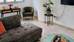 Ein modernes Wohnzimmer mit einem grauen Sofa, einem kleinen runden Tisch mit Korb und Würfel, einem grünen Topfpflanzen und einem grauen Sessel, sowie einem an der Wand montierten Fernseher.