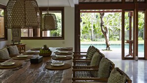 A spacious room with a wooden dining table set for a meal, surrounded by woven chairs and a view of a lush garden and pool outside.