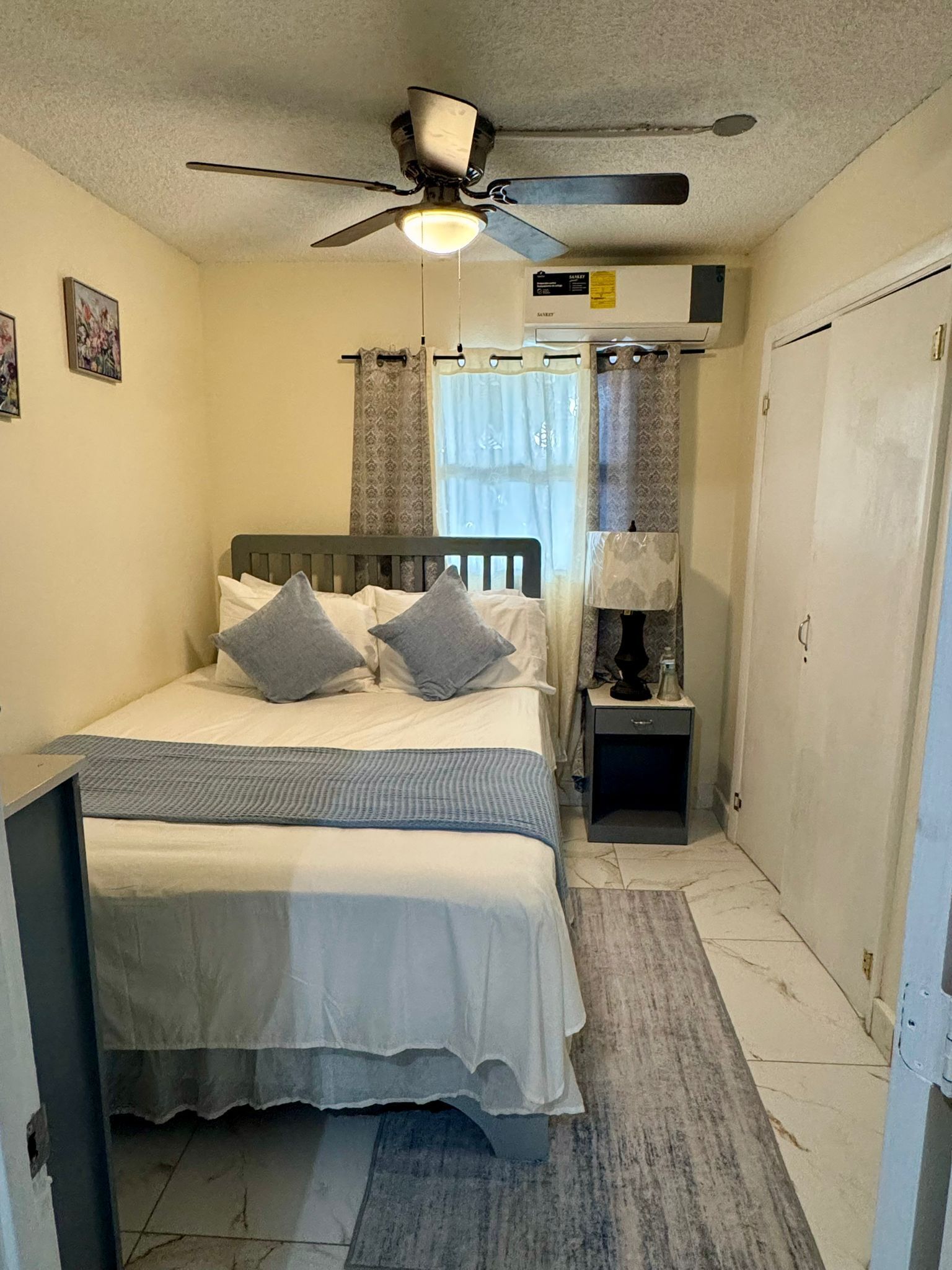 A small, neatly arranged bedroom with a single bed, a bedside table, a nightstand, a ceiling fan, and a window with curtains.