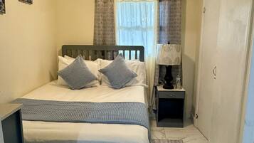A small, neatly arranged bedroom with a single bed, a bedside table, a nightstand, a ceiling fan, and a window with curtains.