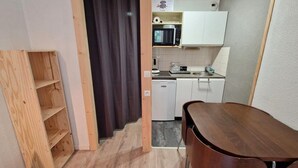 A compact kitchen with white cabinets, a microwave, and a sink. There's a wooden shelf unit and a dark curtain near the entrance.