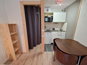 Cozinha compacta com armários brancos, micro-ondas e pia. Há uma estante de madeira e uma cortina escura perto da entrada.