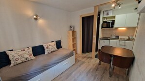A compact room with a bed, a dining table, a kitchen area, and a built-in shelf.