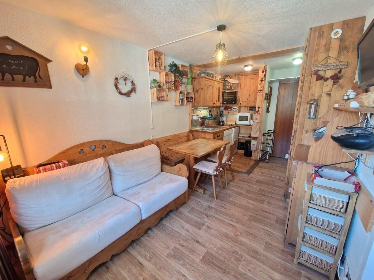Un salon chaleureux avec un canapé, une table à manger en bois et des chaises. On aperçoit, au fond, une cuisine avec des meubles en bois et un luminaire suspendu.
