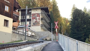 A railway crossing with a pedestrian path, a building with a mural, and a forested background.