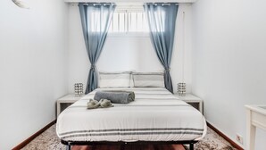 Un dormitorio con una cama, dos mesitas de noche, una ventana con cortinas y una lámpara de noche pequeña.