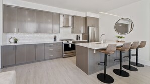 A modern kitchen with grey cabinets, a central island with bar stools, stainless steel appliances, and a round mirror on the wall. - The Theodore 2BD/2B - All King Bedrooms (New Orleans)
