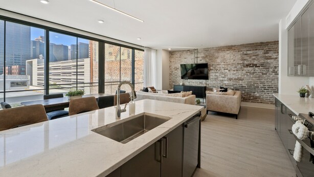 A modern kitchen with a large island, a dining area with chairs, a sofa, and a brick wall. - The Theodore 2BD/2B - All King Bedrooms (New Orleans)