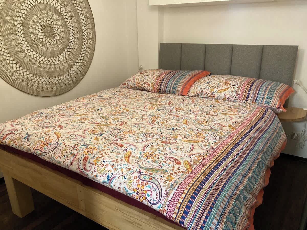 A bed with a patterned bedspread and pillows, a wooden headboard, and a wall hanging.