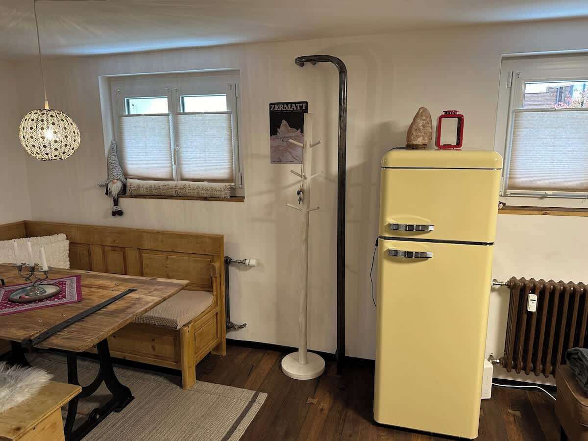 Ein kleiner Raum mit einer Holzbänkl, einem Tisch mit Unterlage, einem gelben Kühlschrank, einer Heizung und einem Fenster mit Rollläden.