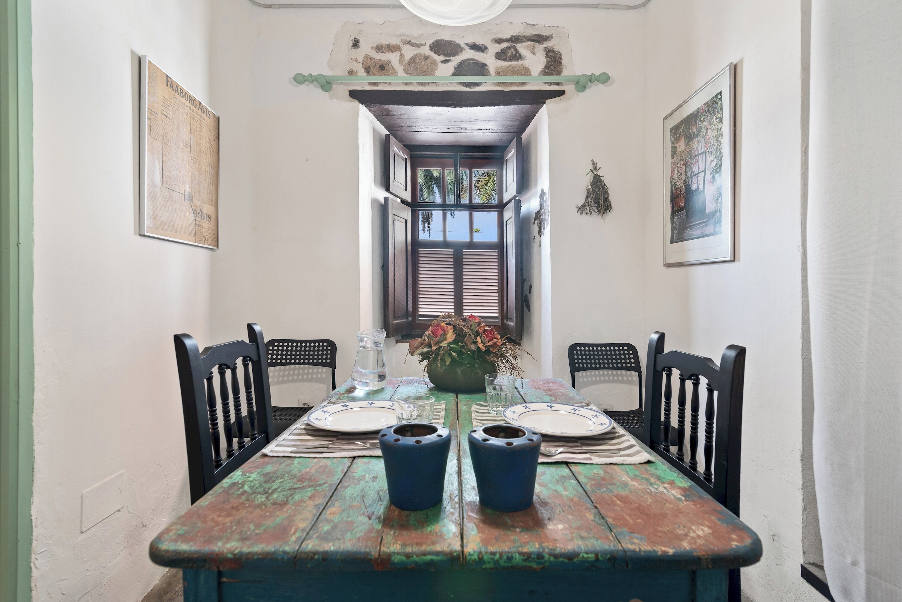 A dining table with two blue cups, a vase with flowers, and a window with shutters.
