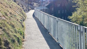 A paved pathway alongside a stone retaining wall, leading to a residential area with houses and a street lamp. - Cozy Zermatt Studio: Terrace, Village Views (Zermatt)