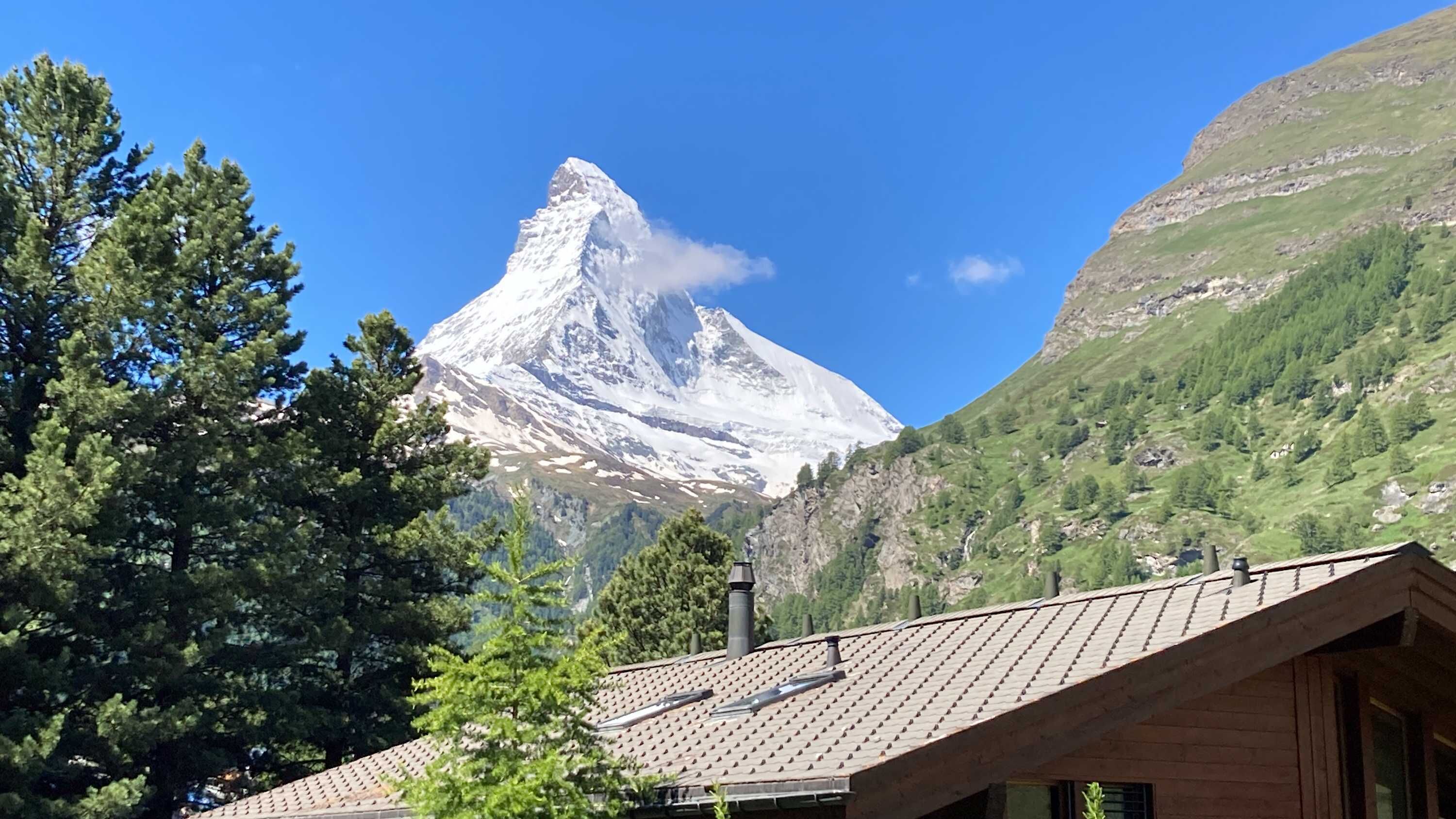 Une montagne dont le sommet est enneigé, un bâtiment en bois avec un toit de tuiles et une végétation luxuriante tout autour.