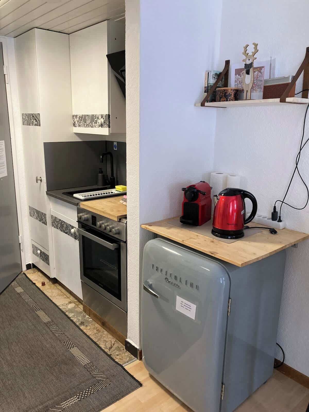 Eine kleine Küche mit eingebautem Backofen, einem kleinen Kühlschrank und einer Kaffeemaschine auf einer Holzausziehtafel.
