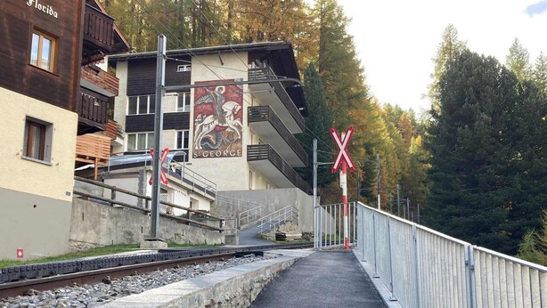 A railway crossing with a pedestrian path, a building with a mural, and a forested background. - Studio Alpine Zermatt: Village views & Terrace (Zermatt)