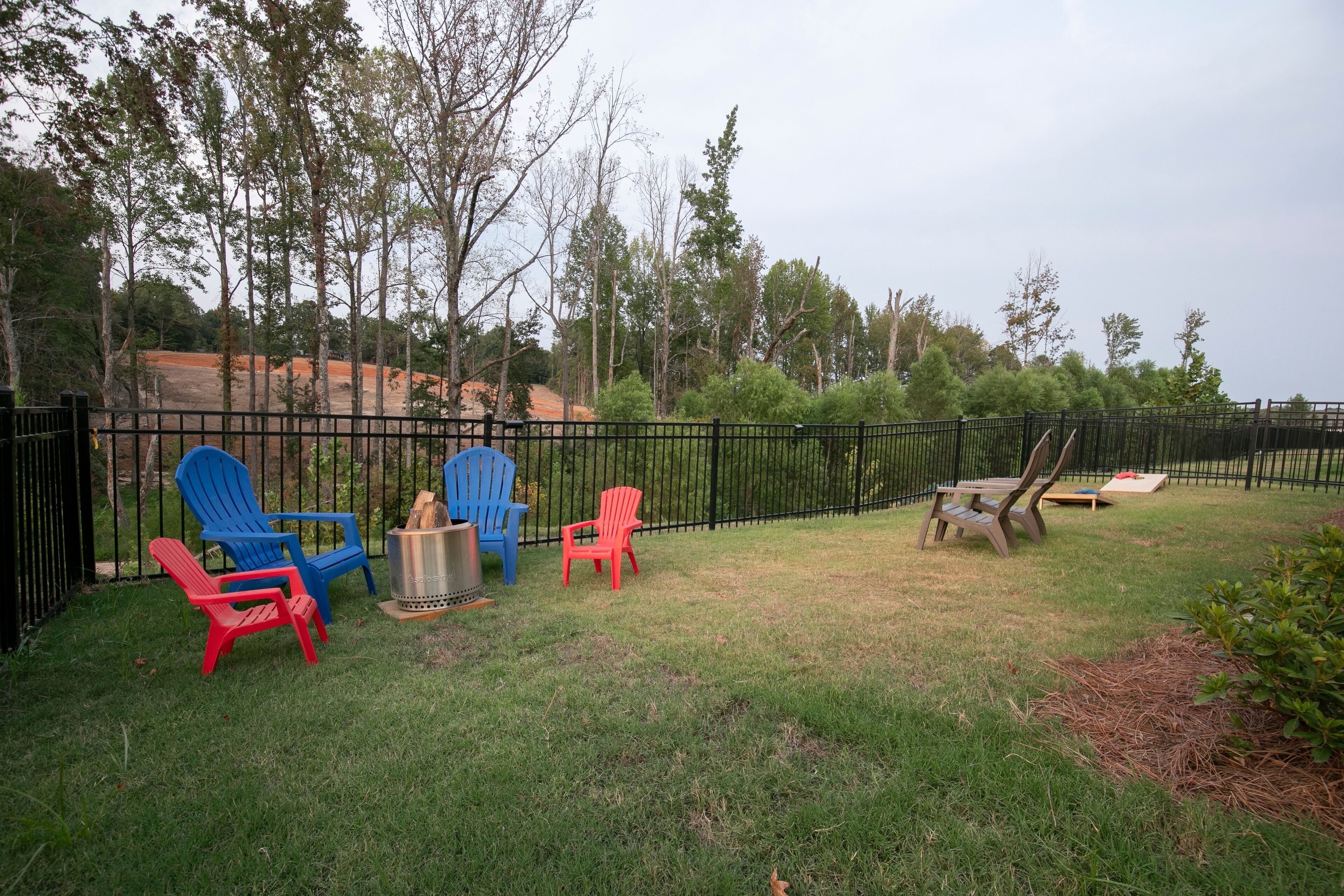 A backyard with a black metal fence, several Adirondack chairs in red and blue, a picnic table, and a small fire pit.