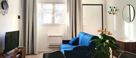 Una sala moderna con un sofá azul, una mesa de comedor con sillas, un televisor sobre un mueble bajo y una planta en maceta.