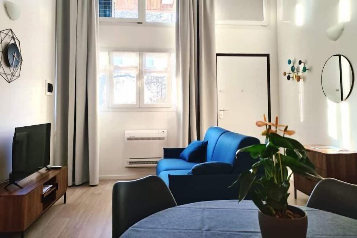 A modern living room with a blue sofa, a TV on a wooden stand, a dining table with chairs, and a potted plant.