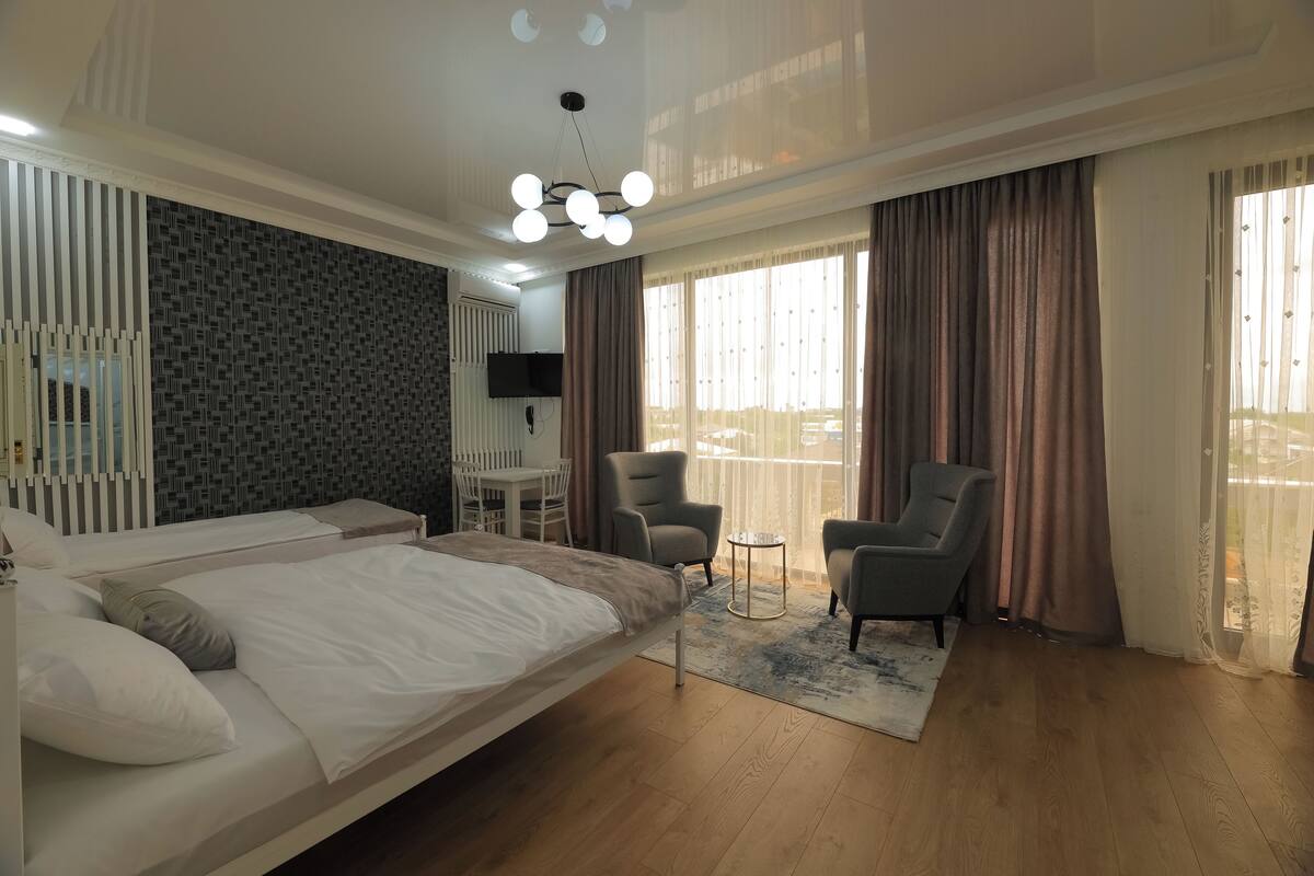 Ein Hotelzimmer mit einem großen Bett, einem Schreibtisch, zwei Sesseln, einem Fernseher und einem Balkon mit Vorhängen.