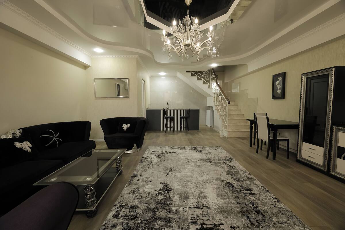 Ein geräumiges Wohnzimmer mit einem modernen Kronleuchter, einem schwarzen Sofa, einem Glastisch und einem gemusterten Teppich.