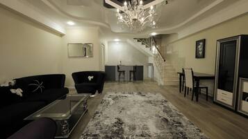 A spacious living room with a modern chandelier, a black sofa, a glass coffee table, and a patterned rug.