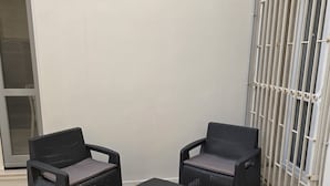 Un patio con dos sillones de mimbre y un otoman a juego, una pared blanca y una reja metálica.