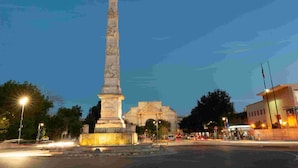 Un monumento alto en una plaza pública, con un edificio al fondo.