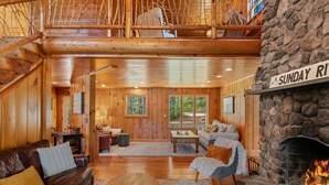 A spacious wooden interior with a stone fireplace labeled "SUNDAY RIVER," a leather sofa, a coffee table, and a view of the outdoors through large windows. - The Epic Sunday River Getaway (Bethel)