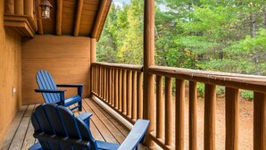A wooden deck with two Adirondack chairs and a table, overlooking a forested area. - The Epic Sunday River Getaway (Bethel)