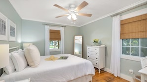 Une chambre à coucher comprenant un lit, une commode, un ventilateur de plafond et deux fenêtres équipées de stores.
