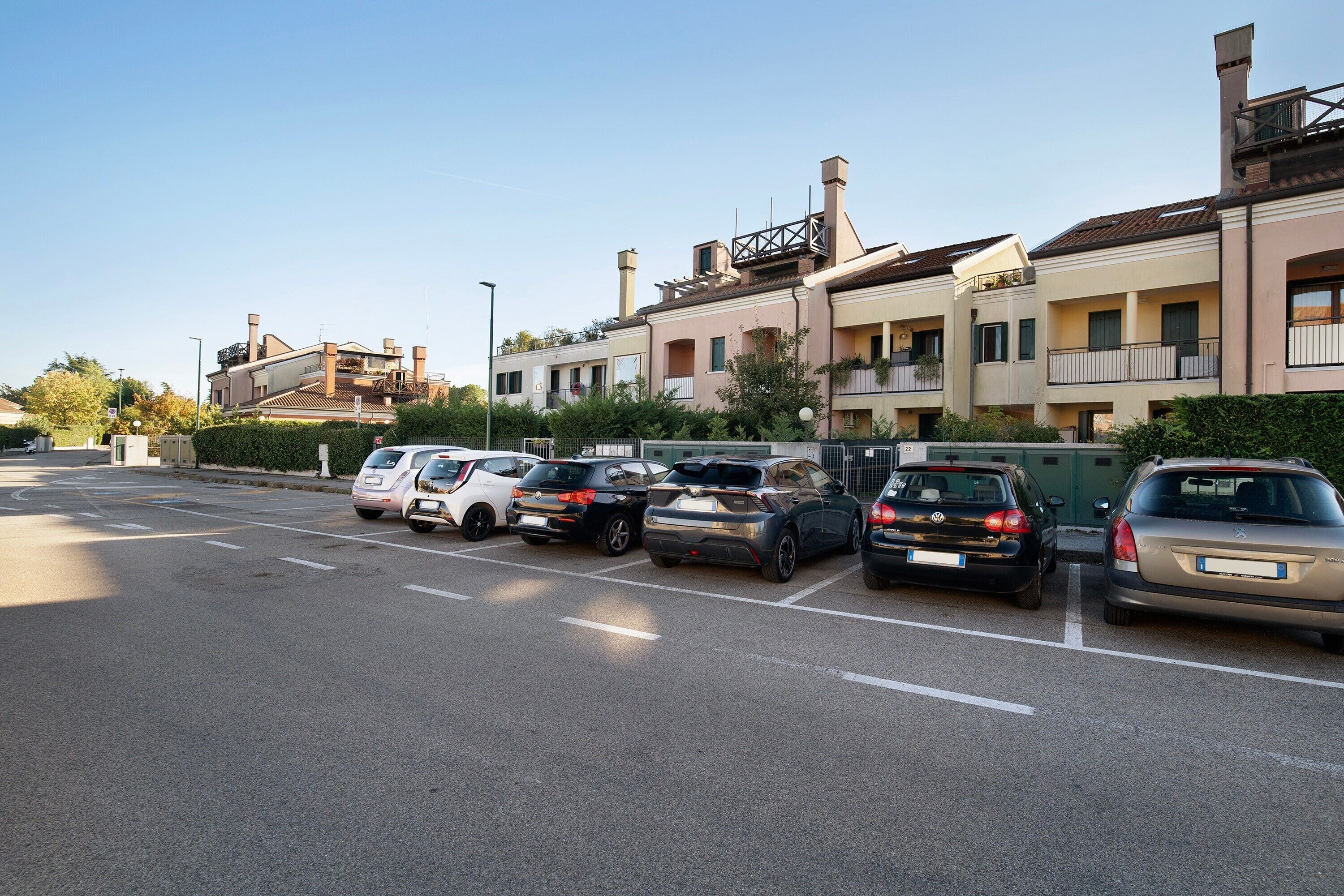 A parking lot with several cars, a clear sky, and residential buildings with balconies.