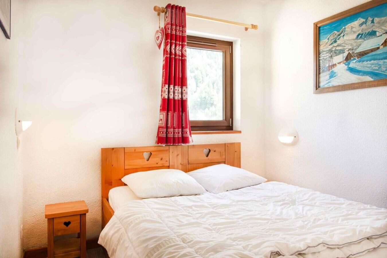 A bedroom with a wooden headboard, a single bed with white bedding, a nightstand, a window with a red curtain, and a painting on the wall.