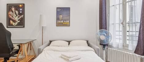 A hotel room with a bed, a desk, a chair, a standing lamp, a poster, a window with curtains, and a radiator.