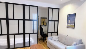 A modern room with a grey sofa, a small wooden table, and a black office chair. A poster is on the wall, and a sliding door with frosted glass panels is visible. - Champs de Mars - Ecole Militaire 1bdr by B'Your Home (Paris)