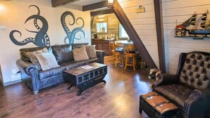 A cozy living room with a leather sofa, a coffee table, and a tufted armchair. A kitchen area is visible in the background. - Pirate 2-story Villa Near Beach (Ocean Shores)