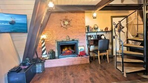 A cozy living room with a brick fireplace, wooden flooring, a bookshelf, and a painting of a ship on the wall. - Pirate 2-story Villa Near Beach (Ocean Shores)