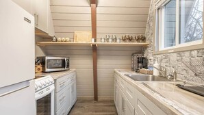 Cocina compacta con muebles blancos, microondas, estufa, repisa de madera con cristalería y protector de piedra.