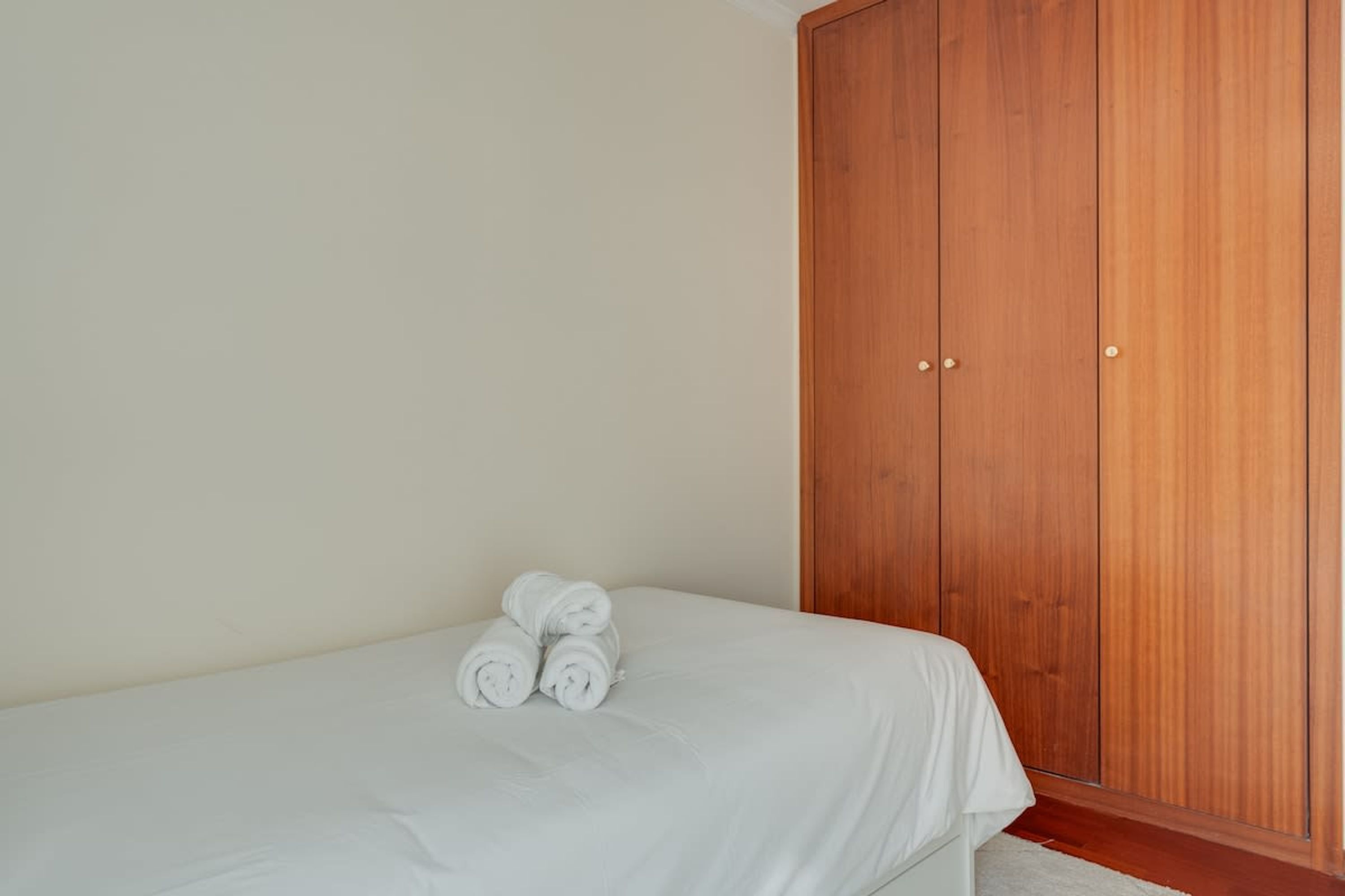 A single bed with white linens and rolled towels, a wooden wardrobe, and a plain wall.