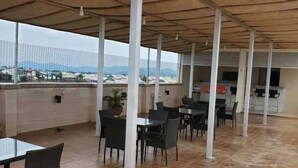A covered outdoor dining area with tables and chairs, a view of the cityscape, and a tiled floor. - TILT APARTMENT (Bwari)