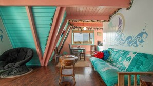 A room with a wooden ceiling, teal walls, a wooden floor, a black chair, a round table, a teal sofa with colorful pillows, and a window with a view. - Hawaiian 2-story Beach Villa (Ocean Shores)