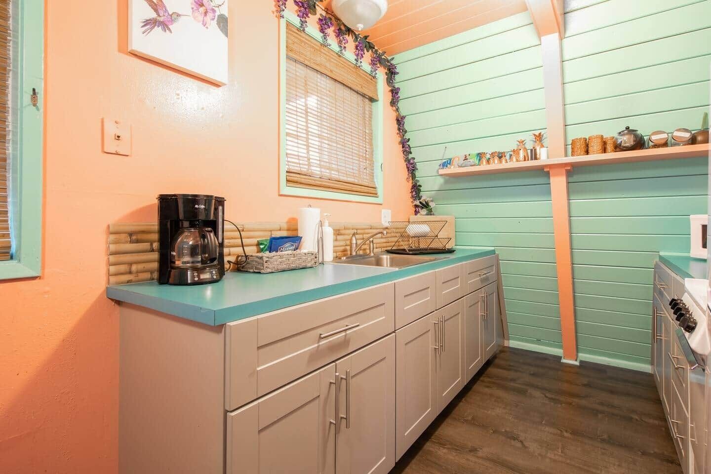 A kitchen with a teal countertop, beige cabinets, a coffee maker, and a window with blinds.