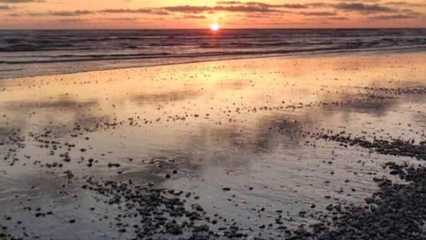 Una playa con el sol poniéndose en el horizonte, reflejado en la arena mojada.