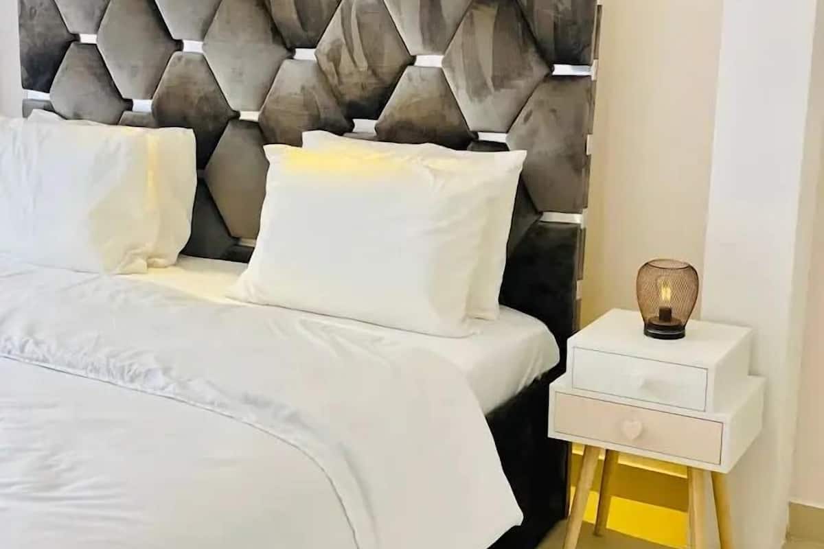A neatly made bed with white bedding and pillows, a modern headboard, and a bedside table with a lamp.