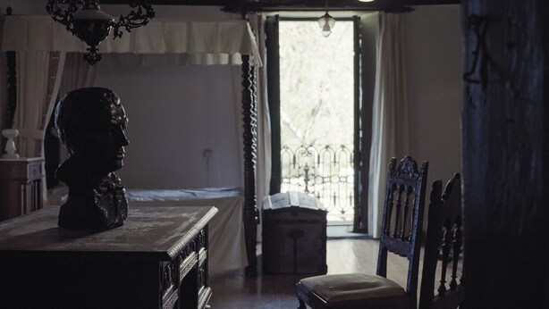 A dimly lit room with a large window, a wooden table with a bust sculpture, and a chair. - A Casa da Memoria - House-museum to live the experience of going back 100 years. (Aldea de Xullán)
