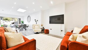 Een moderne woonkamer met oranje banken, een wit vloerkleed, een eethoek met stoelen en een aan de muur bevestigde televisie.