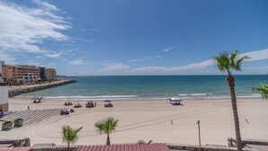 A beach with a clear view of the ocean, a few palm trees, and a building with multiple balconies. - Las Gaviotas 436 Mid-century Ocean View Apartment (Puerto Peñasco)