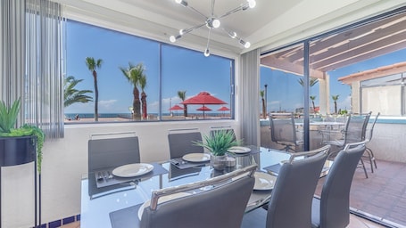 A dining area with a view of the beach, featuring a glass table, chairs, and red umbrellas.