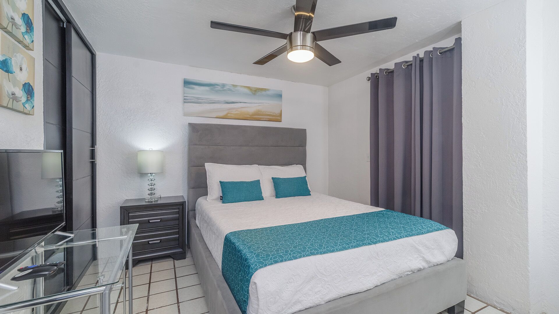 Une chambre à coucher comprenant un lit, un bureau, une chaise, un ventilateur de plafond et une fenêtre avec des rideaux.