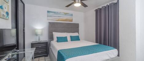Une chambre à coucher comprenant un lit, un bureau, une chaise, un ventilateur de plafond et une fenêtre avec des rideaux.