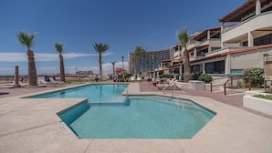 A pool area with two swimming pools, lounge chairs, palm trees, and a building with balconies. - Las Gaviotas 436 Mid-century Ocean View Apartment (Puerto Peñasco)